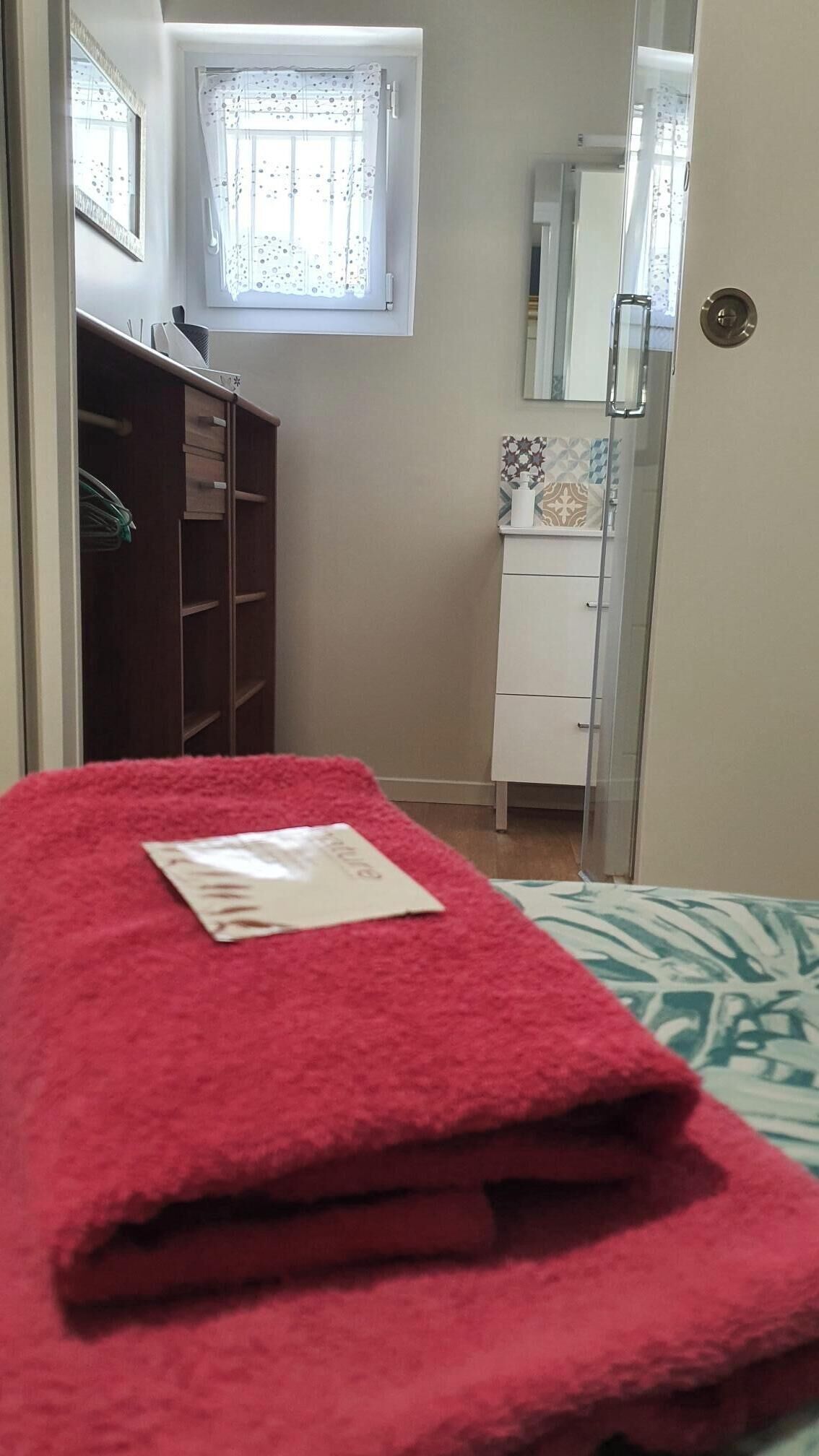 Ein Schlafzimmer mit einem roten Handtuch auf dem Bett, einem weißen Nachttisch, einem Kleiderschrank, einem kleinen Fenster mit Vorhang und einem Spiegel an der Wand.