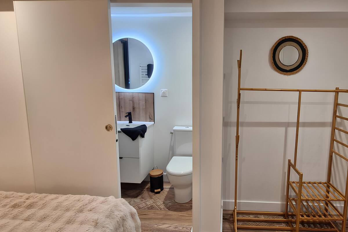 A modern bathroom with a toilet, sink, and mirror. There is a wooden rack and a window with a view of foliage.
