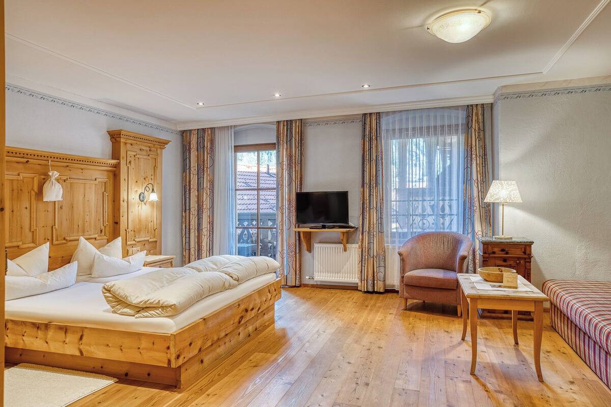 A wooden bed with pillows, a television, a chair, a table, and a sofa in a room with wooden flooring and curtains.