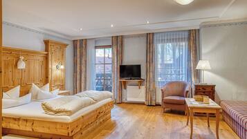 A wooden bed with pillows, a television, a chair, a table, and a sofa in a room with wooden flooring and curtains.