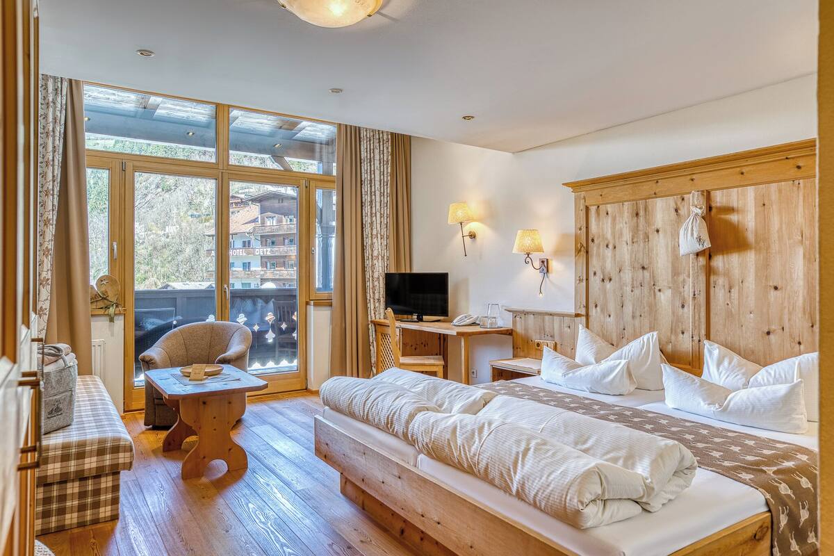 A hotel room with a large bed, a sofa, a wooden table, a TV, and a balcony with a view of buildings.