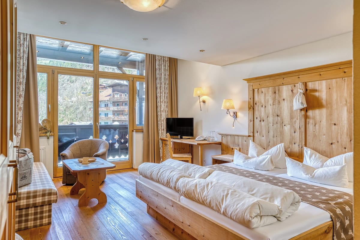 Habitación de hotel con una cama grande, un sofá, una mesa de madera, un televisor y un balcón con vistas a edificios.