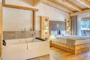 A bedroom with a wooden headboard, a large window, a bathtub, and a bedside table. - Posthotel Kassl (Oetz)