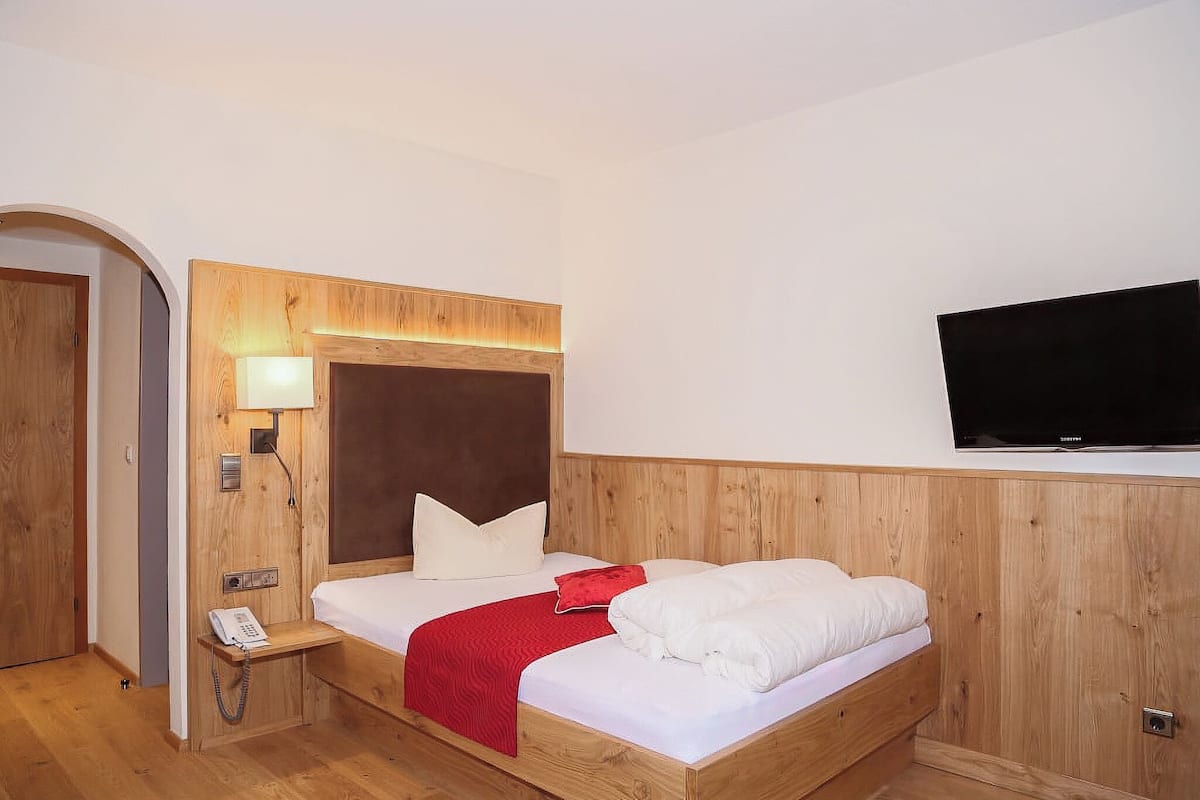 A hotel room with a wooden headboard, a bed with white bedding, a red throw blanket, a bedside lamp, a phone, and a flat-screen TV mounted on the wall.
