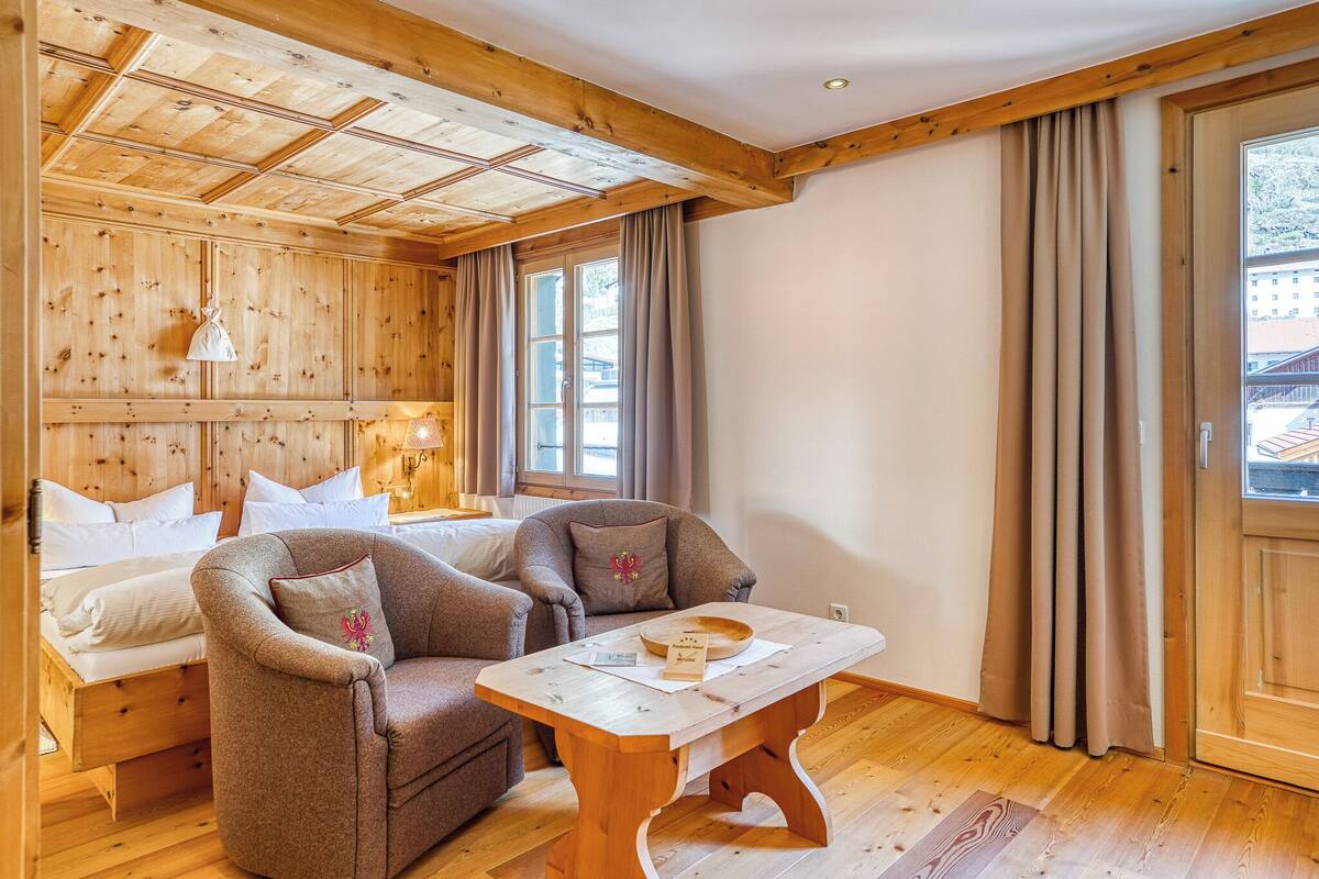 A wooden interior with a bed, two armchairs, a wooden table, and a window with curtains.