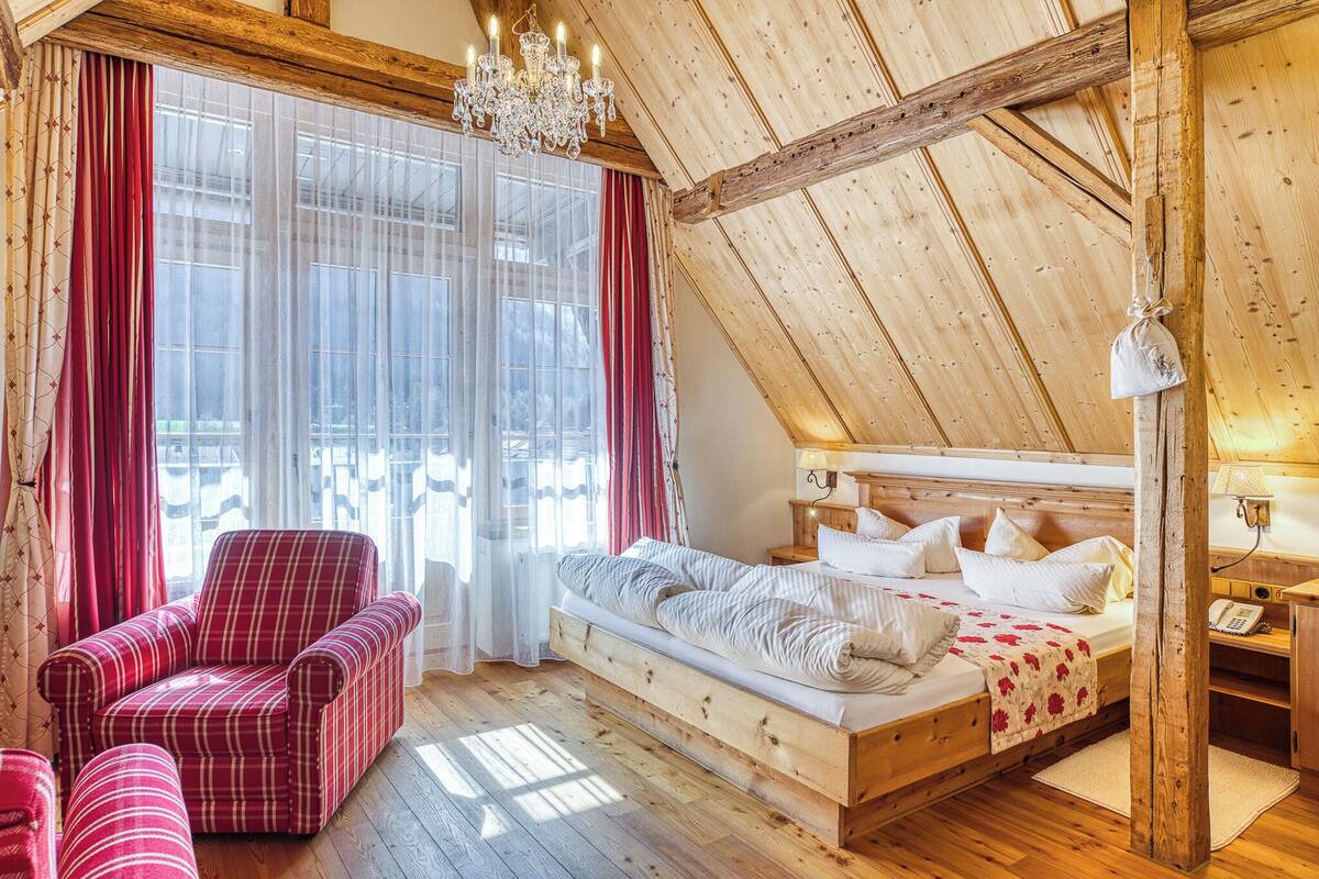 A bedroom with a sloped ceiling, a bed with white bedding, a red plaid armchair, and a chandelier.