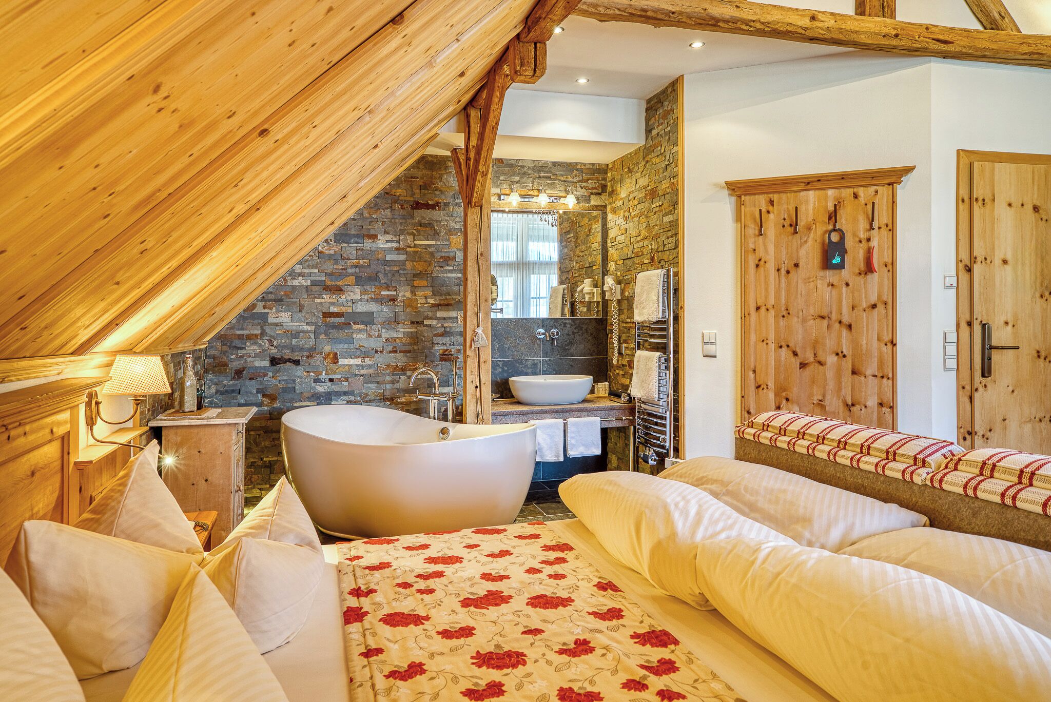 A bedroom with a large bed, a wooden ceiling, a stone wall, a bathtub, and a wooden wardrobe.