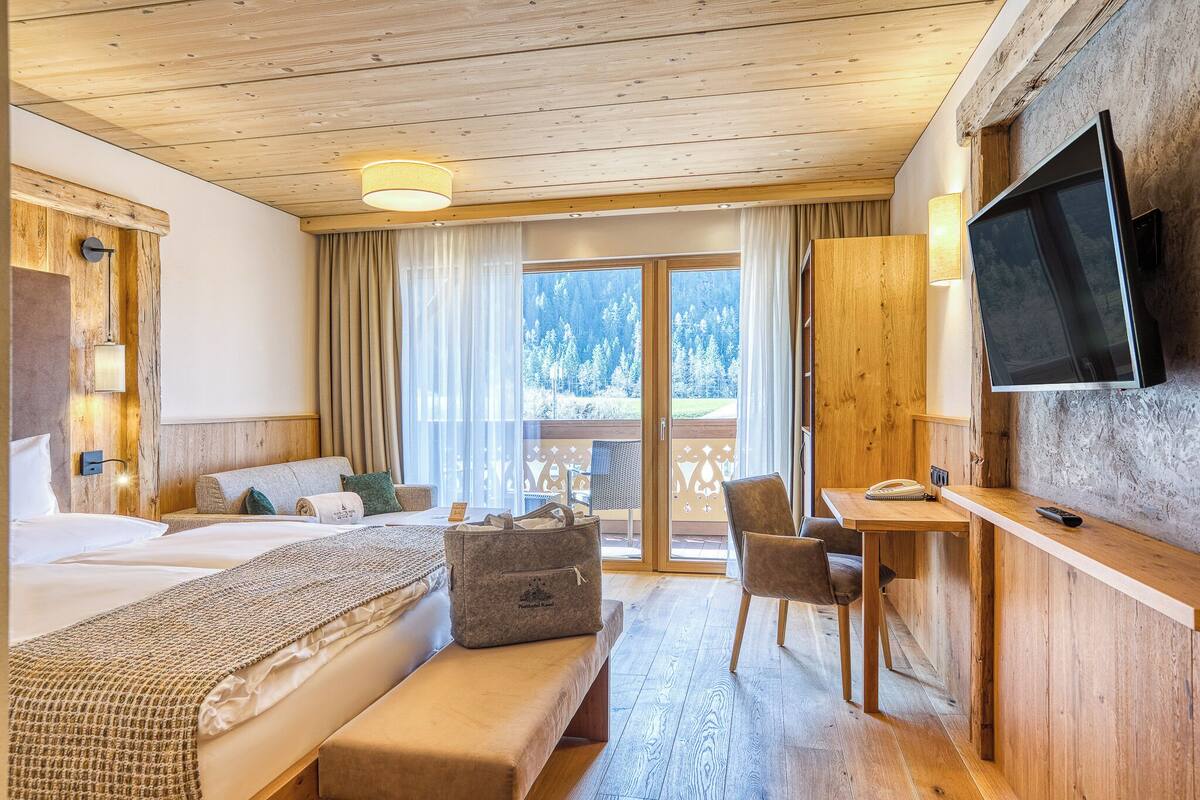 A hotel room with a wooden ceiling, a bed with a bench, a desk with a chair, a television, and a view of a snowy forest.