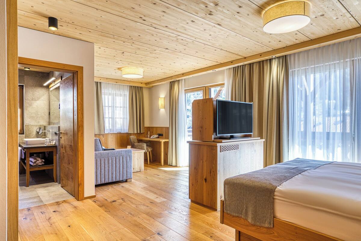 A hotel room with a wooden floor, a bed, a TV, a desk, and a bathroom with a mirror and sink.