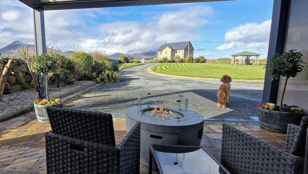 An outdoor patio with wicker furniture, a fire pit, and a view of a house and mountains. - Gleann Ciuin (Newry)