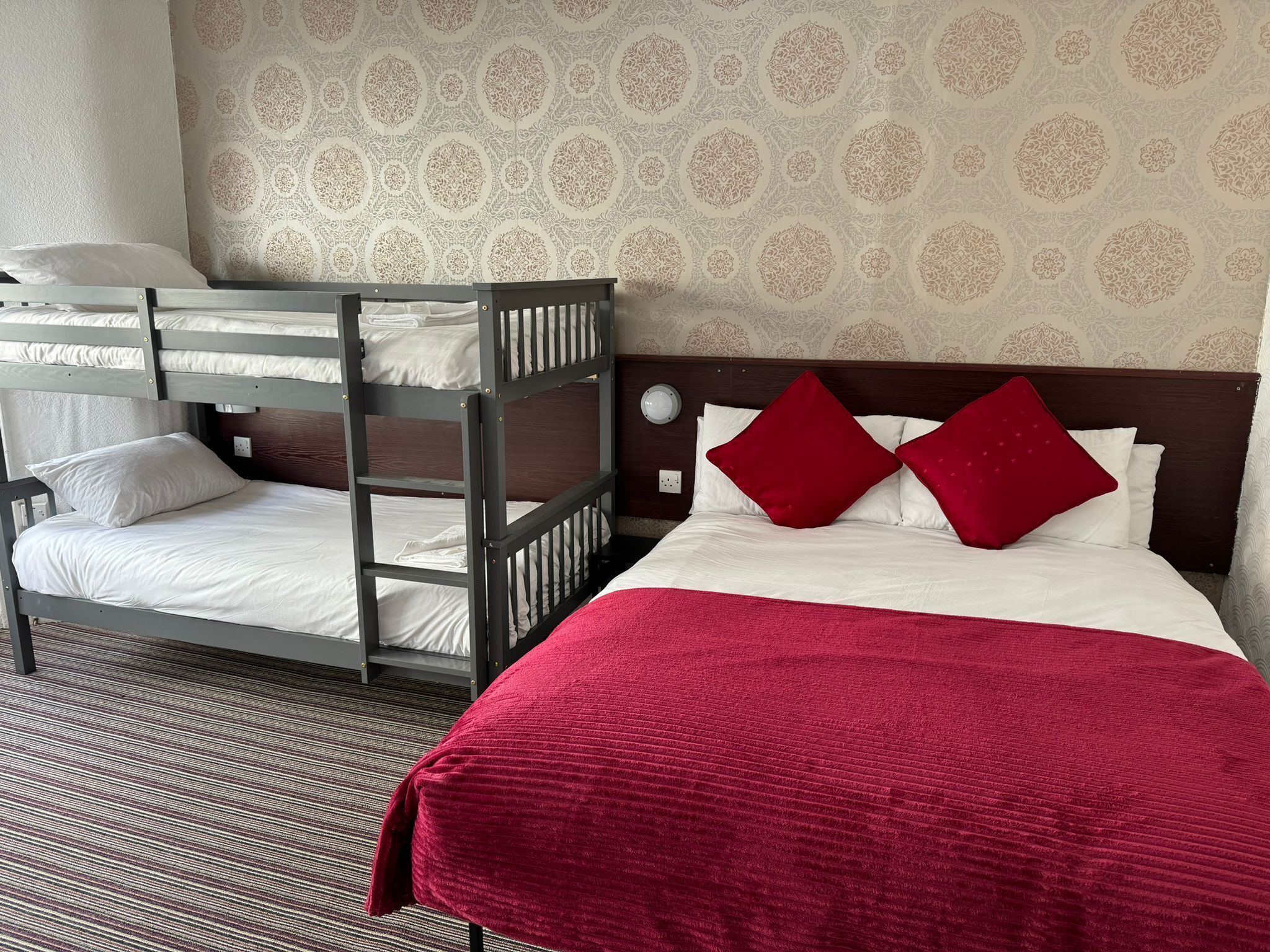 Une chambre d’hôtel avec un lit superposé, un grand lit avec des oreillers rouges et un papier peint à motifs.