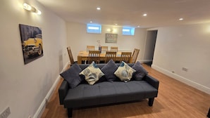 Uma sala de estar moderna com um sofá cinza escuro, mesa de jantar de madeira e cadeiras. Há um quadro na parede e uma pequena janela acima da mesa.