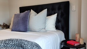 A neatly made bed with a black headboard, multiple pillows, a grey knitted blanket, and a small black side table with books and candles. - Enfield Lux- Brand New Apartment (Auckland)