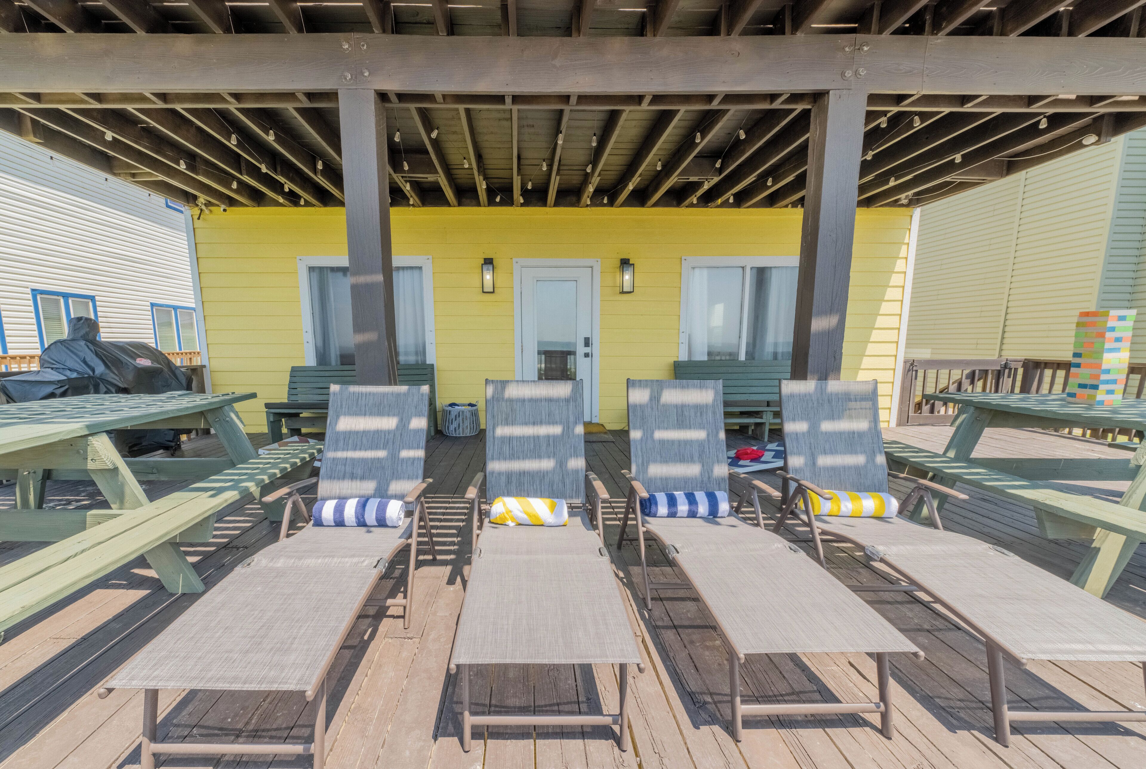 Een overdekt terras met houten banken en loungestoelen, met op de achtergrond een geel gebouw en een houten terras.