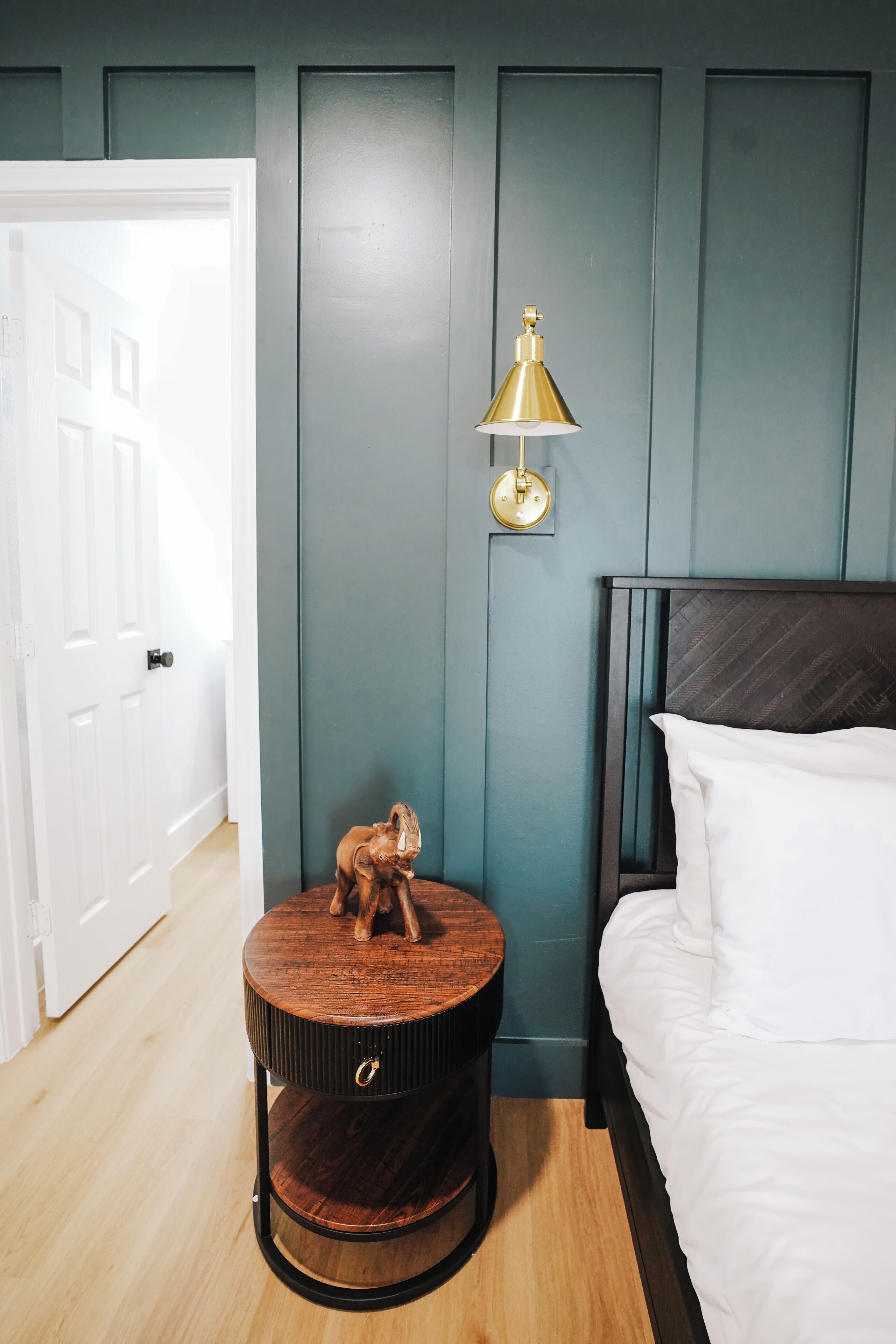 Un dormitorio con una cama, una mesita de noche de madera redonda con una pequeña figurita, y una lámpara de pared.