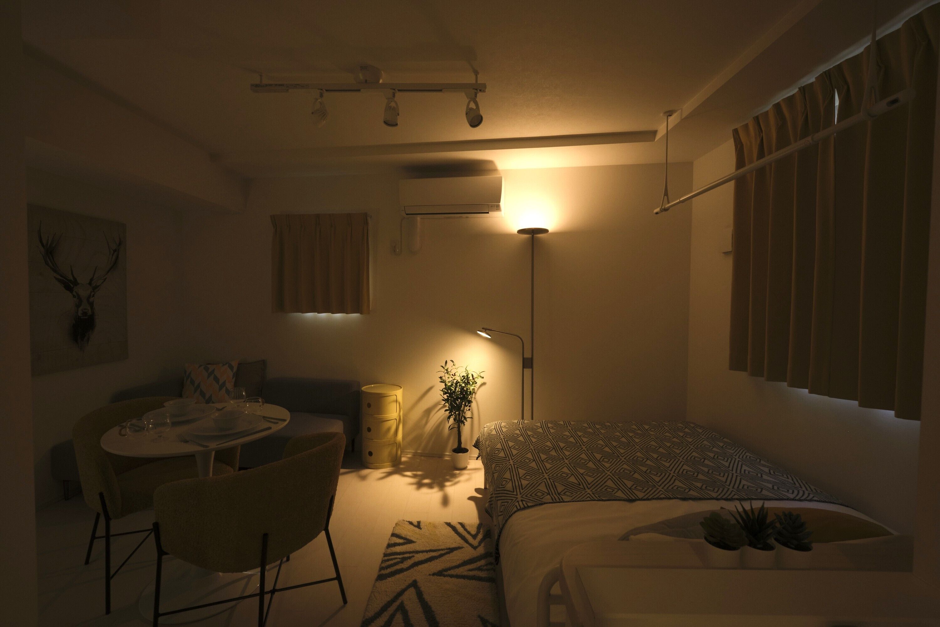 A compact room with a bed, a dining table, a chair, a lamp, and a wall-mounted air conditioner.