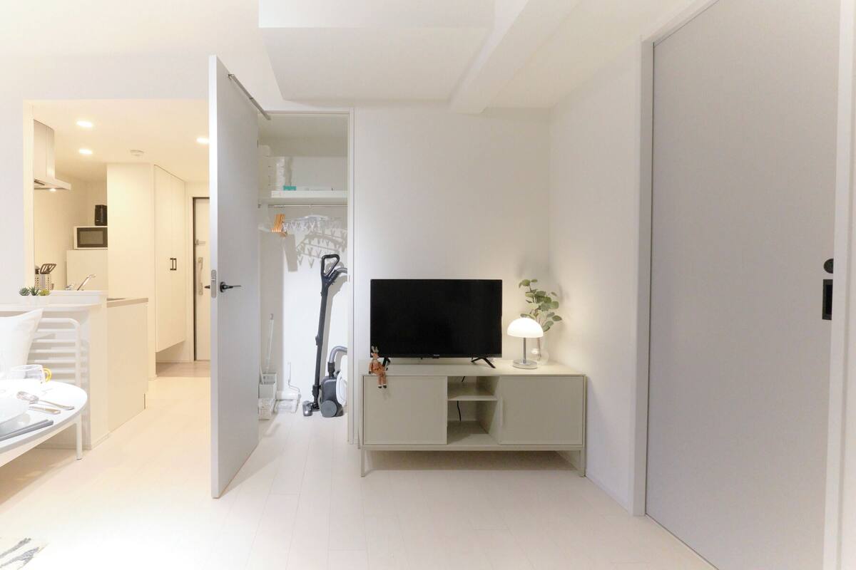 Un salon moderne avec un téléviseur posé sur un meuble blanc, un coin repas avec une table et des chaises, et une cuisine en arrière-plan.