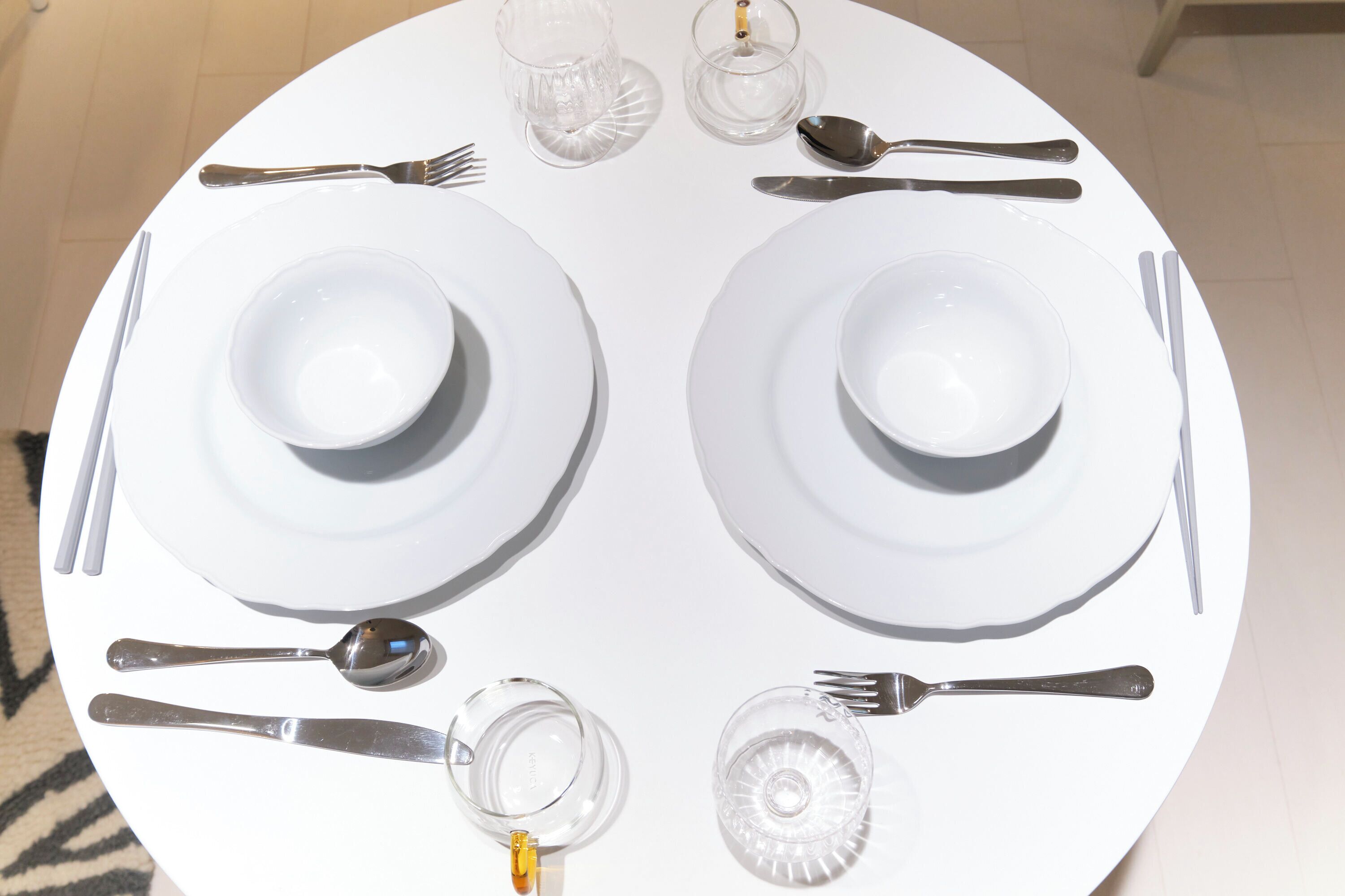A round table set for two with white plates, cutlery, and clear glassware.