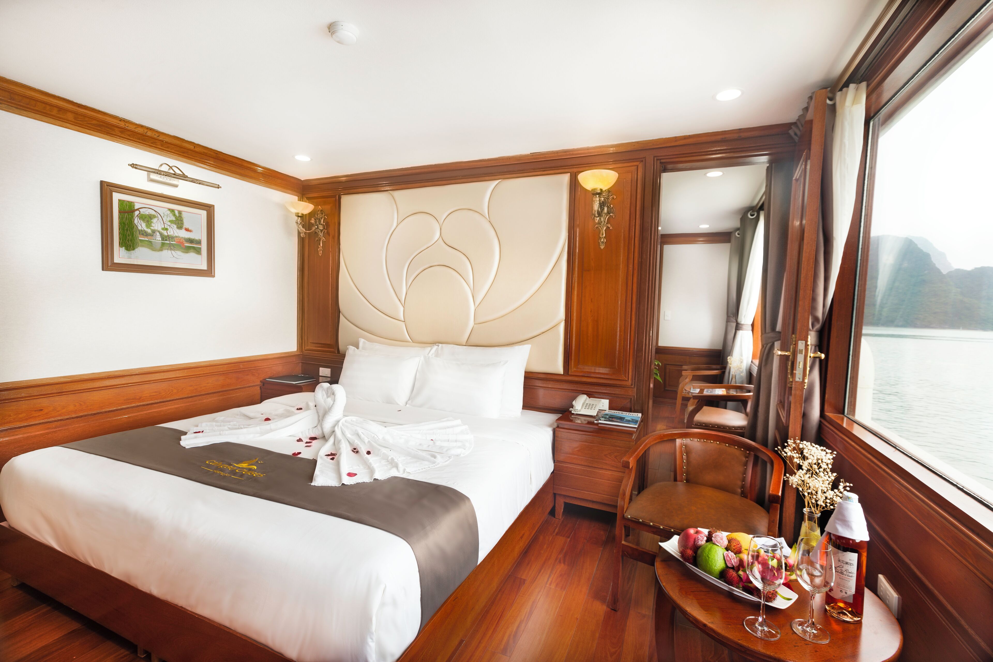 A hotel room with a large bed, a bedside table with a lamp, a chair, a small table with a fruit platter and glasses, and a window with a view of the water.