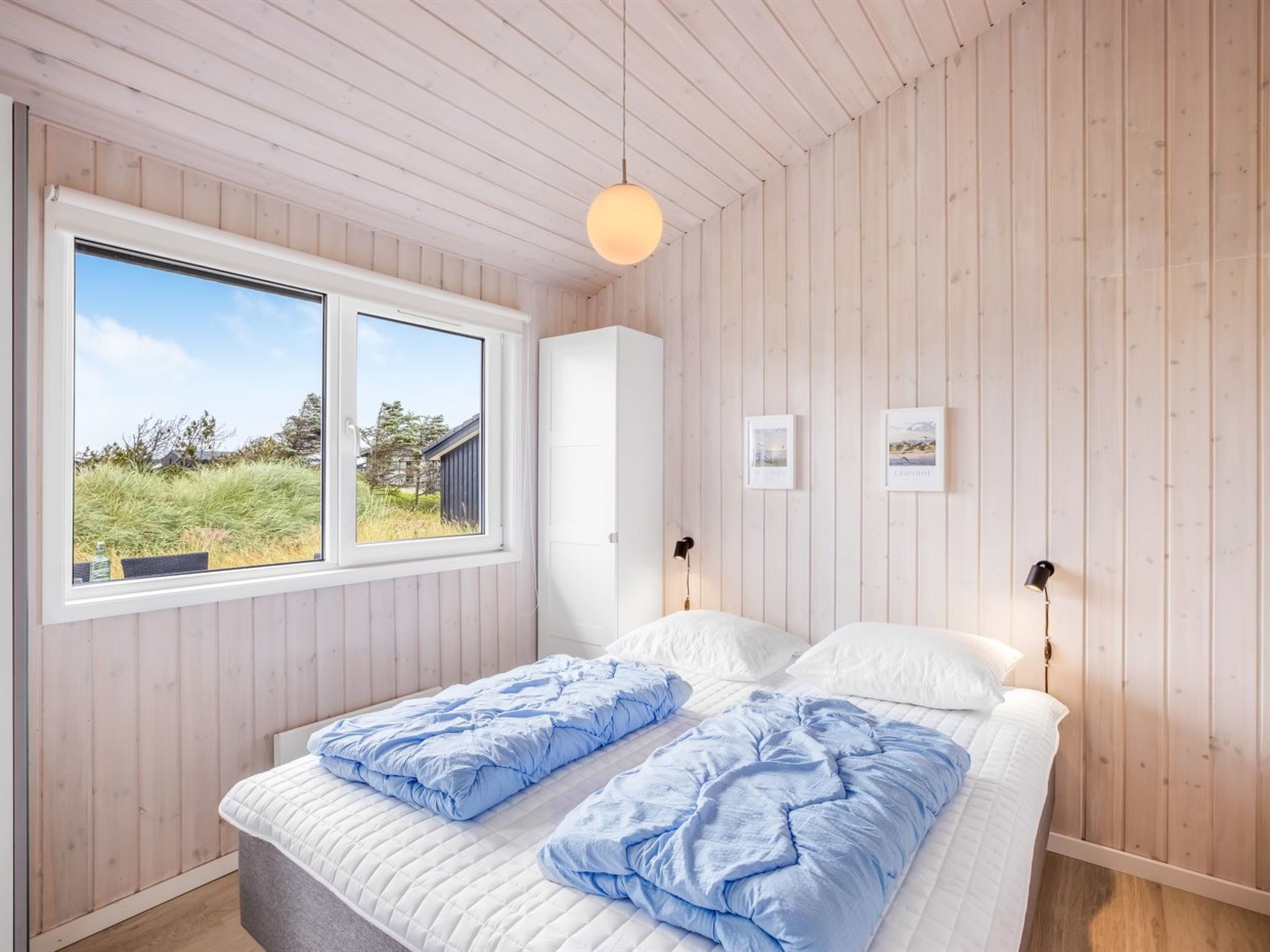 A bedroom with a large window, wooden walls, a bed with blue and white bedding, and two wall-mounted lamps.