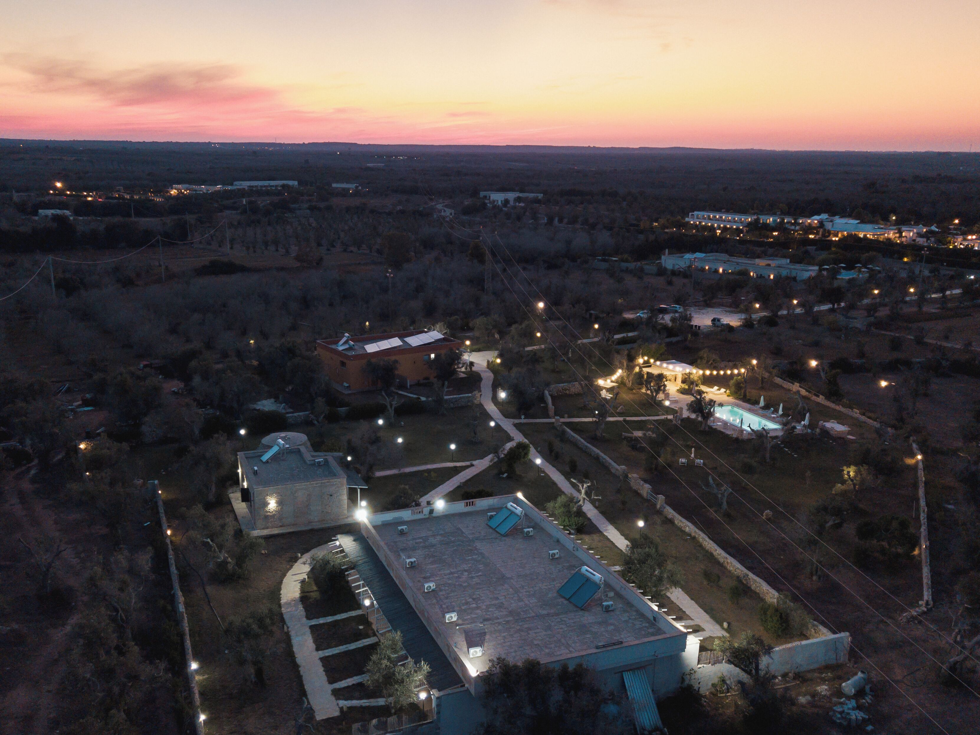 An aerial view of a resort with multiple buildings, a swimming pool, and a well-lit landscape at dusk.