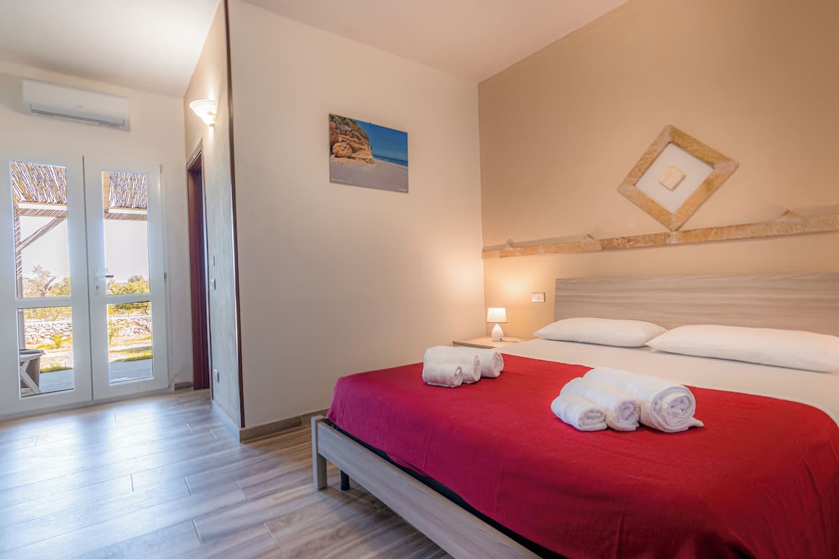 A hotel room with a bed, a red bedspread, a nightstand with a lamp, and a picture on the wall.