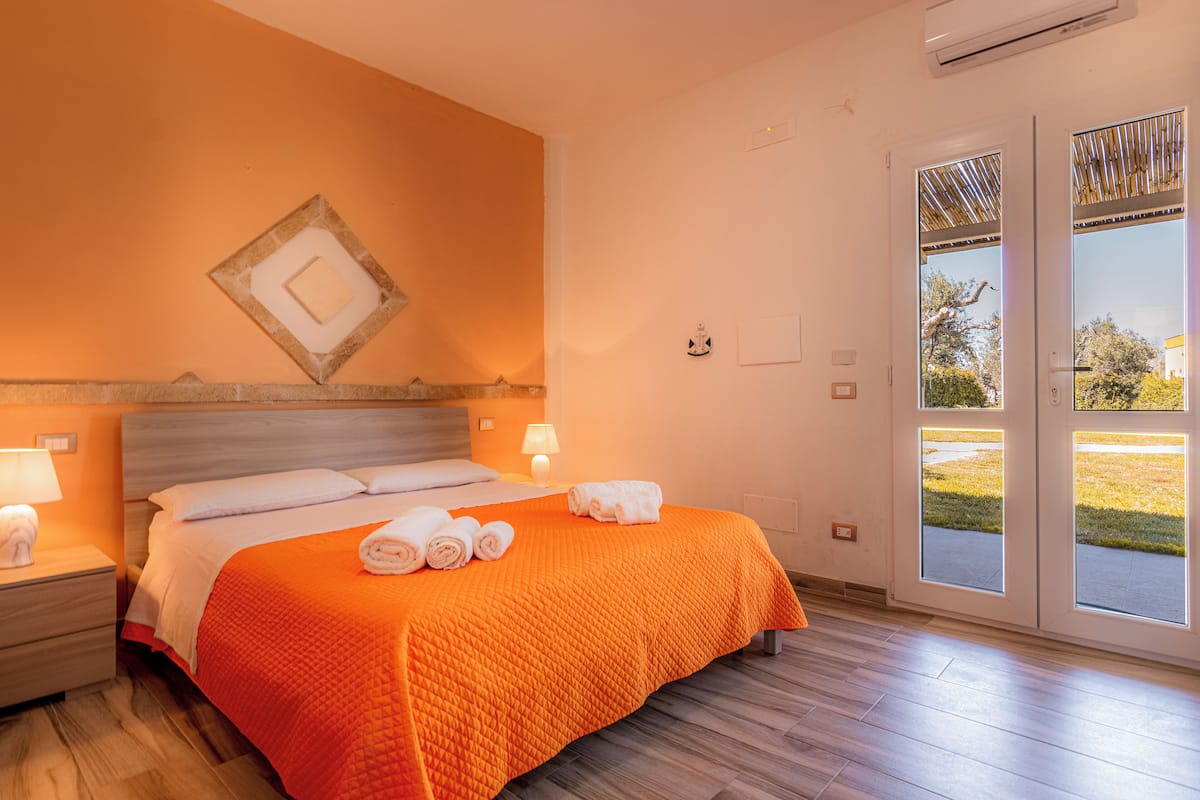 A hotel room with a large bed, a nightstand with a lamp, and a view of the outdoors through a glass door.