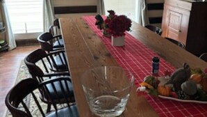 Un comedor con una mesa de madera larga, sillas, un florero con flores y un mantel a cuadros rojos.