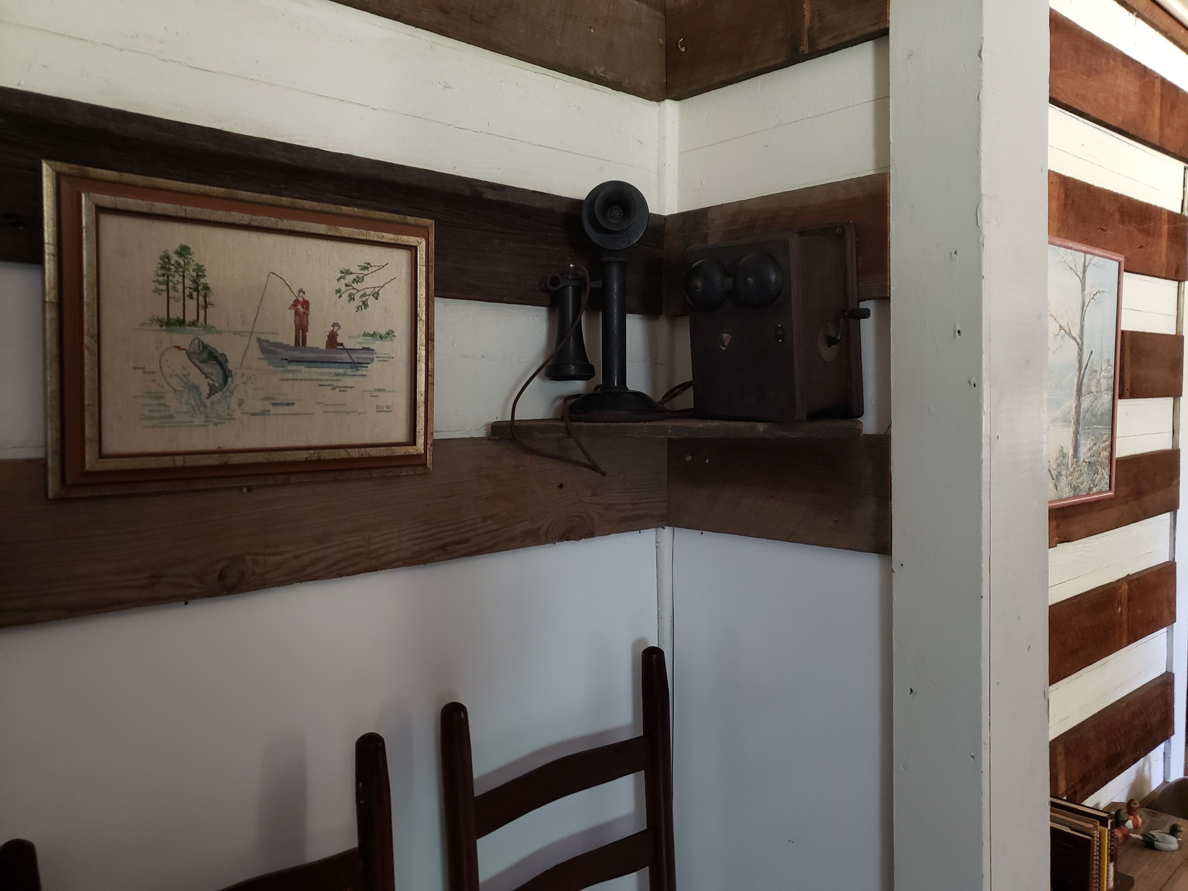 Un coin de pièce avec une chaise en bois, un tableau encadré représentant une scène de pêche et un appareil photo ancien posé sur une étagère.