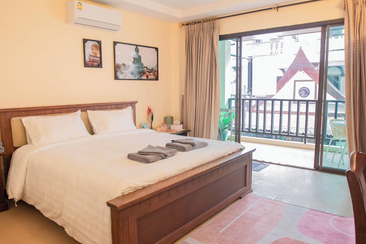 Hotelkamer met een groot bed, nachtkastje, raam met uitzicht op een gebouw en een balkon met stoelen.