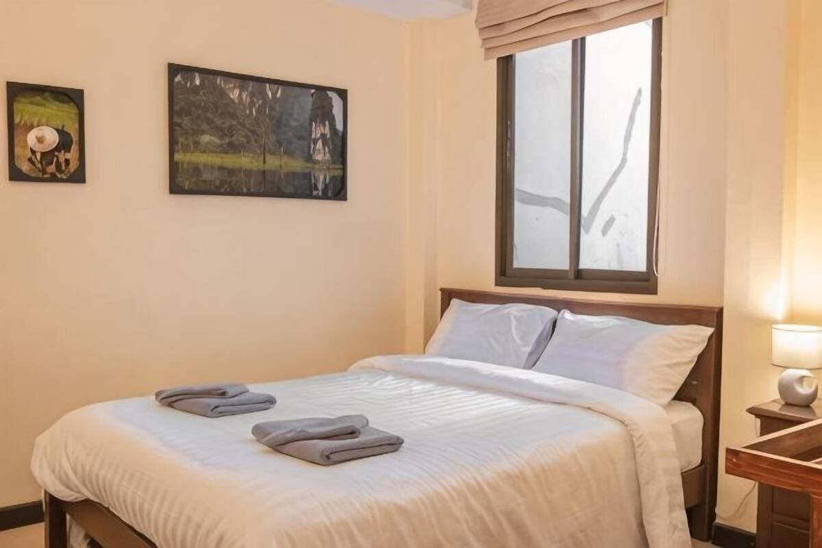 Een net opgemaakt bed met kussens en handdoeken, een raam met gordijnen en twee aan de muur hangende schilderijen.