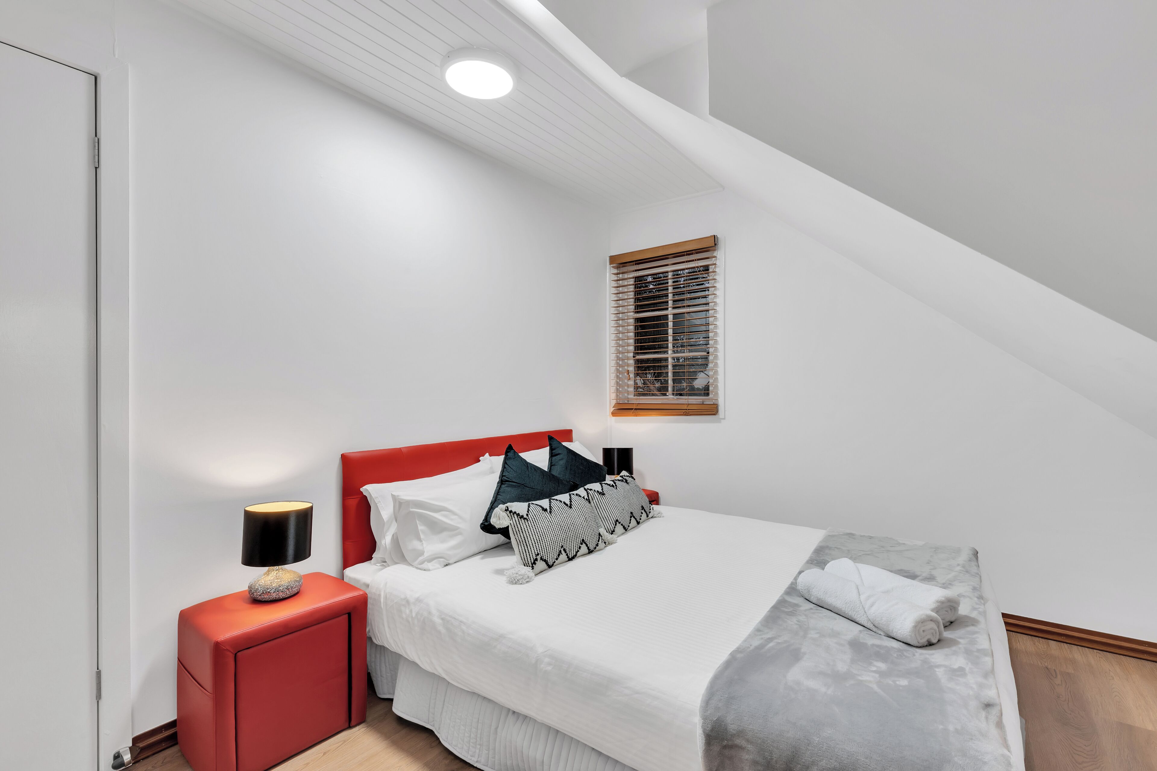 A bedroom with a bed, a red nightstand, a lamp, and a window with blinds.