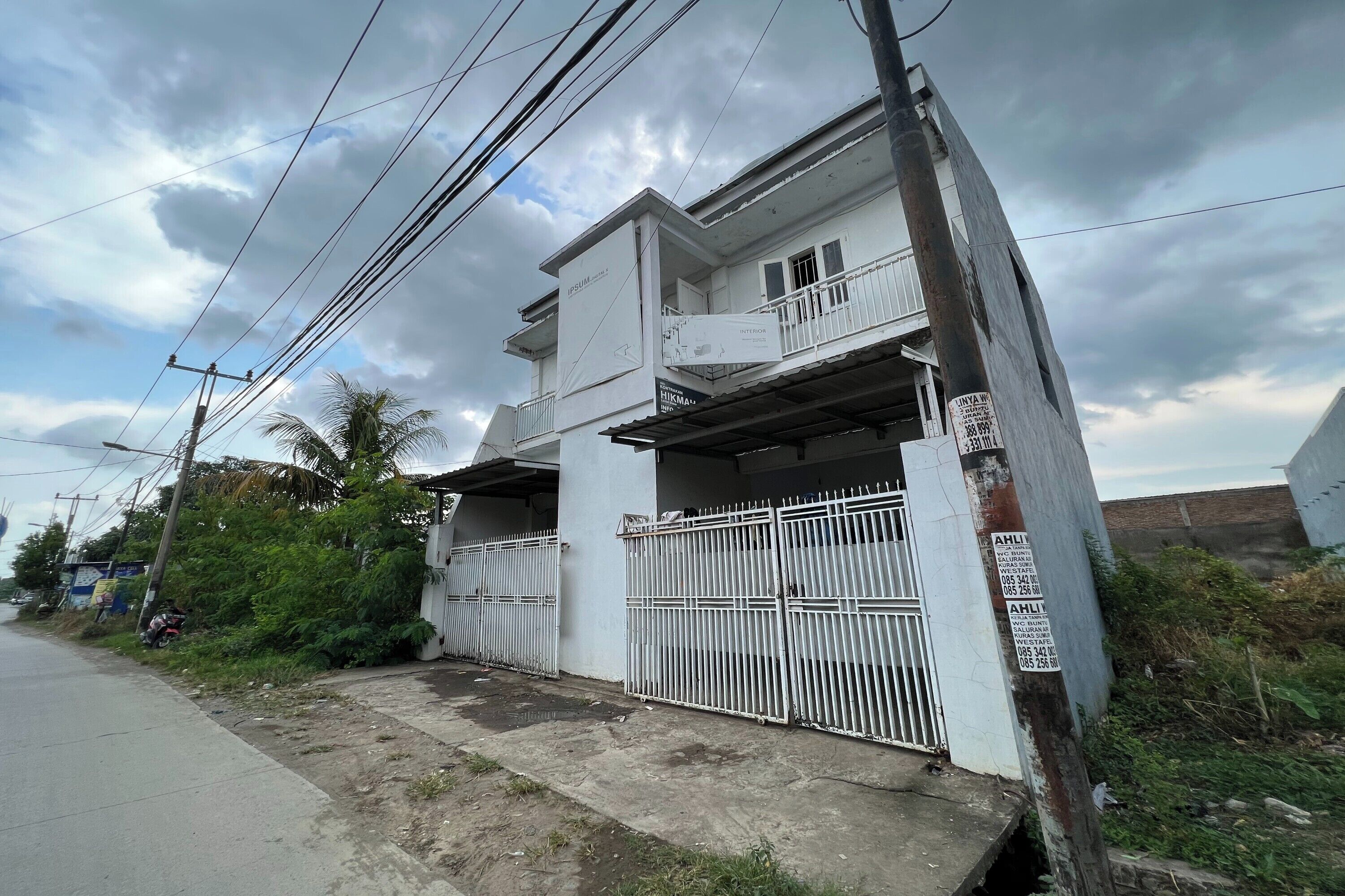 A two-story residential building with a balcony, surrounded by a white metal fence, and utility poles with overhead wires.