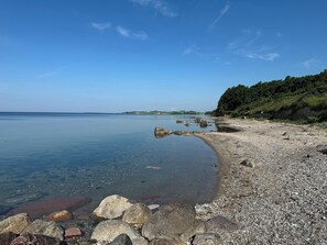 A clear beach with pebbles and rocks, calm water, and a forested hillside.