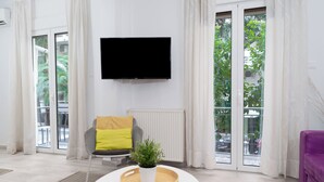 Ein modernes Wohnzimmer mit einem an der Wand befestigten Flachbildfernseher, einem grauen Sessel mit gelbem Kissen, einer kleinen Topfpflanze auf einem weißen Tisch und einem violetten Sofa.