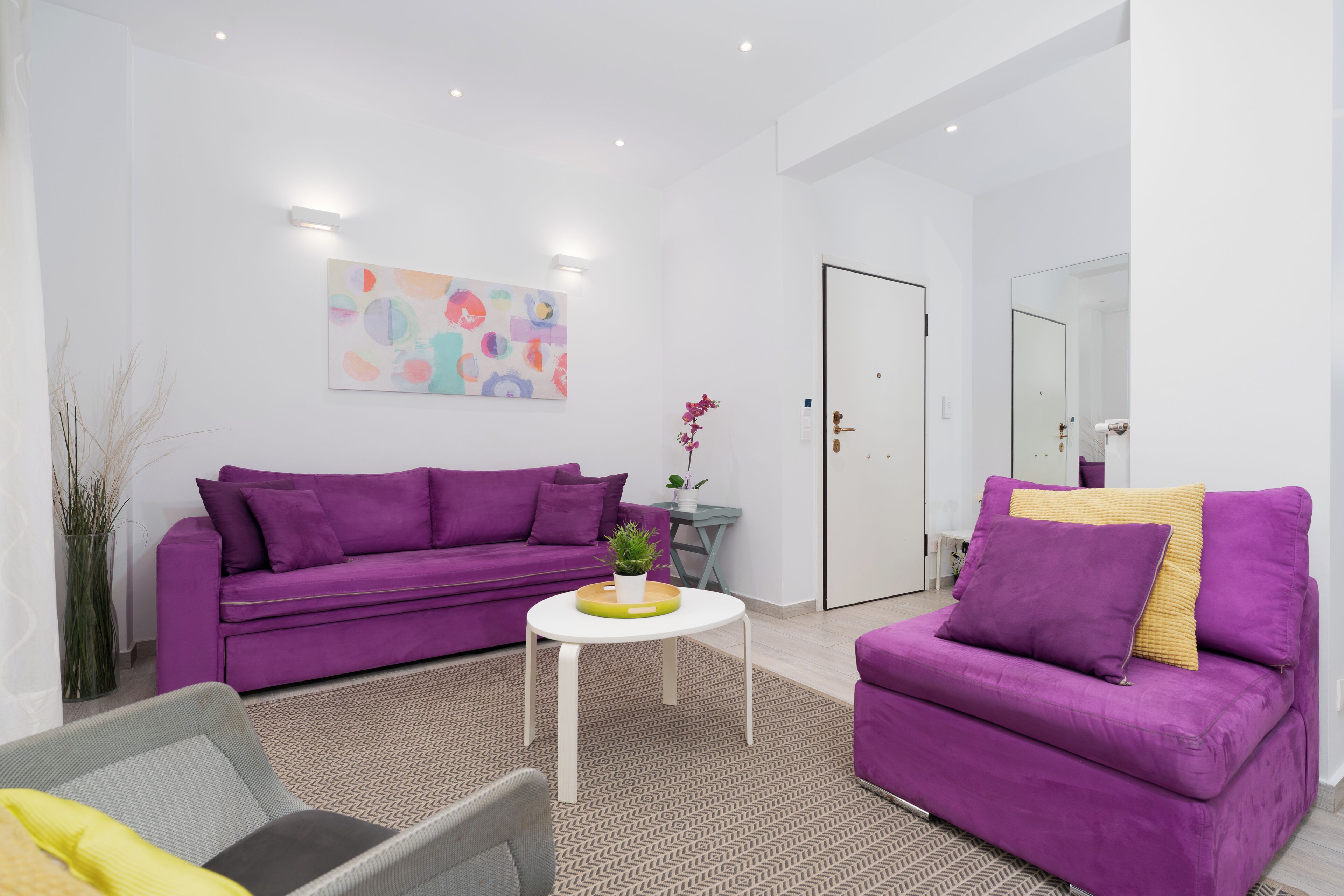 A modern living room with a purple sofa set, a small round table with a plant, and a colorful abstract painting on the wall.