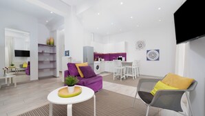 Ein modernes Wohnzimmer mit einem violetten Sofa, einem grauen Sessel, einem kleinen Tisch mit Pflanze und einem Küchenbereich im Hintergrund.