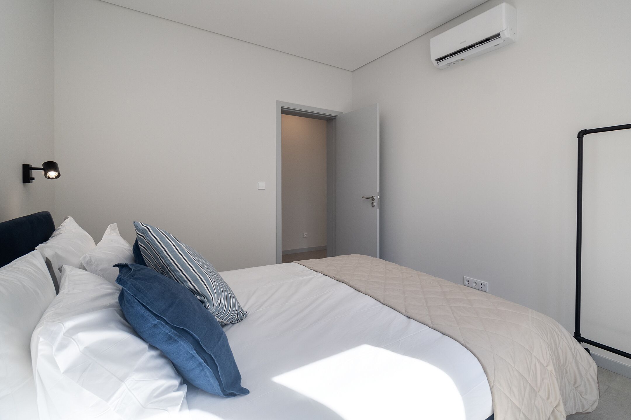 Un dormitorio con cama, almohadas, una lámpara de noche, aire acondicionado y una puerta que da a otra habitación.