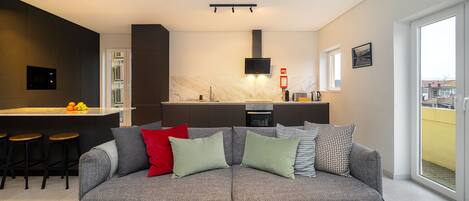 A modern living room with a grey sofa, a kitchen area with a marble countertop, and a large window offering a view.