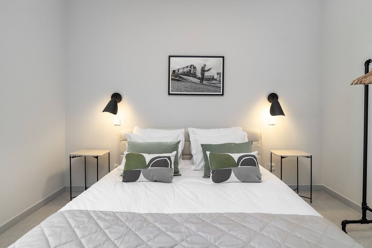 A neatly made bed with white bedding and green and gray pillows, flanked by two black bedside tables with lamps, in a room with a framed picture on the wall.
