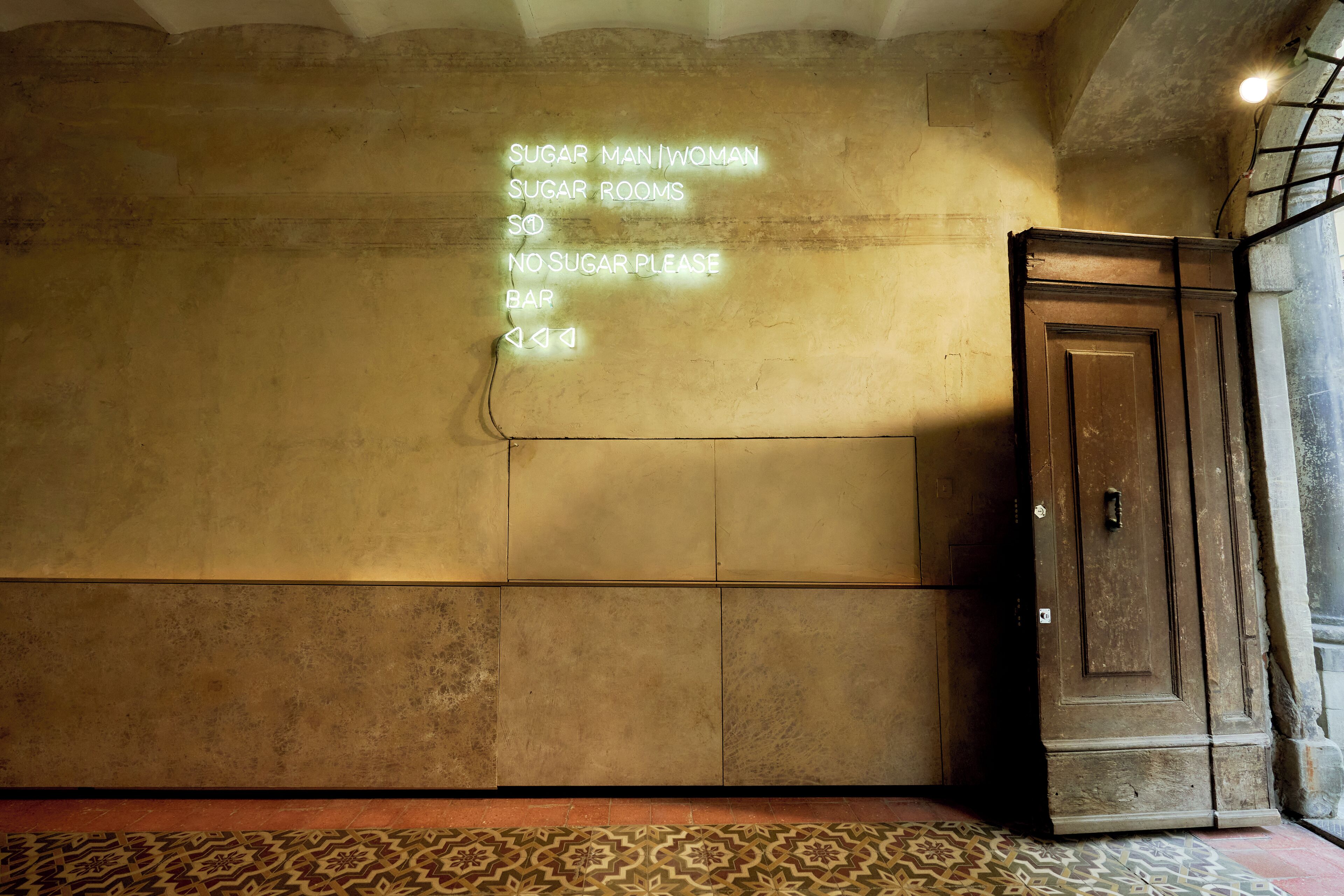 A wall with a neon sign listing amenities: "SUGAR MAN/WOMAN, SUGAR ROOMS, 50, NO SUGAR PLEASE, BAR."