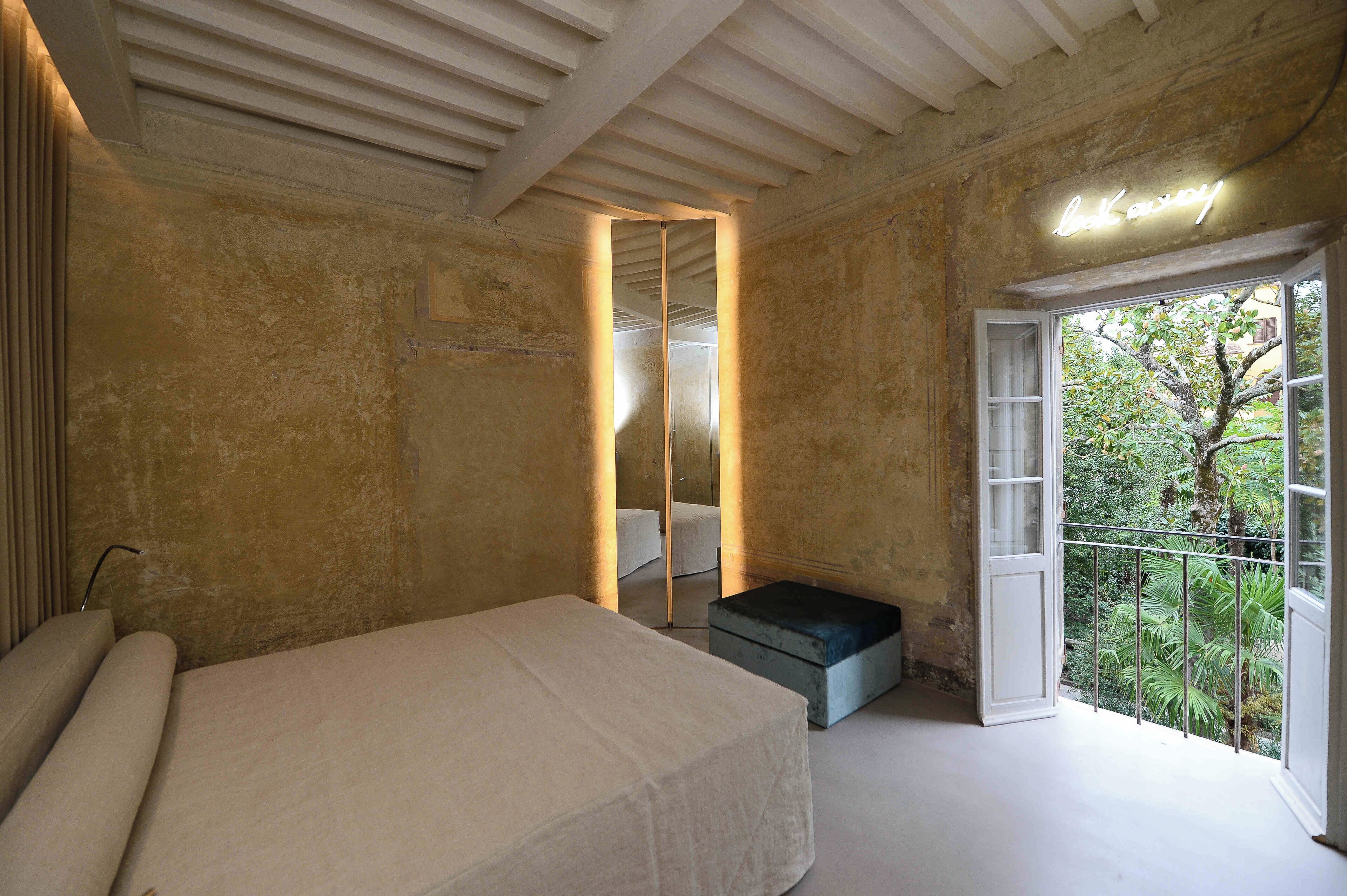 A bedroom with a large bed, a small bench, a mirror, and a balcony with a view of greenery.