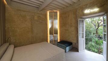 A bedroom with a large bed, a small bench, a mirror, and a balcony with a view of greenery.