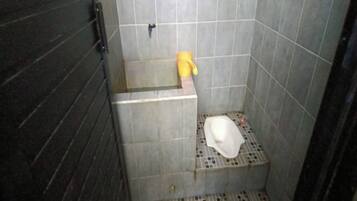 A small, tiled bathroom with a toilet, a yellow bucket, and a metal grate on the floor.