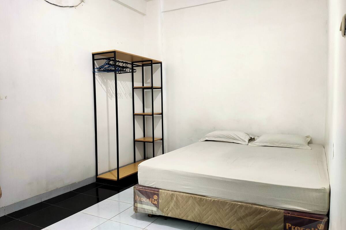 Ein kleines, sauberes Zimmer mit einem Einzelbett, einem schwarzen Metallschrank und einer weißen Wand.