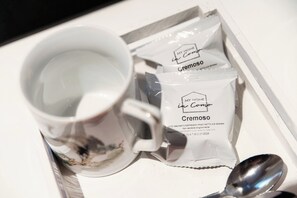 A white mug filled with a clear liquid, possibly water, next to a tray containing two packets of 'Cremoso' coffee and a spoon.