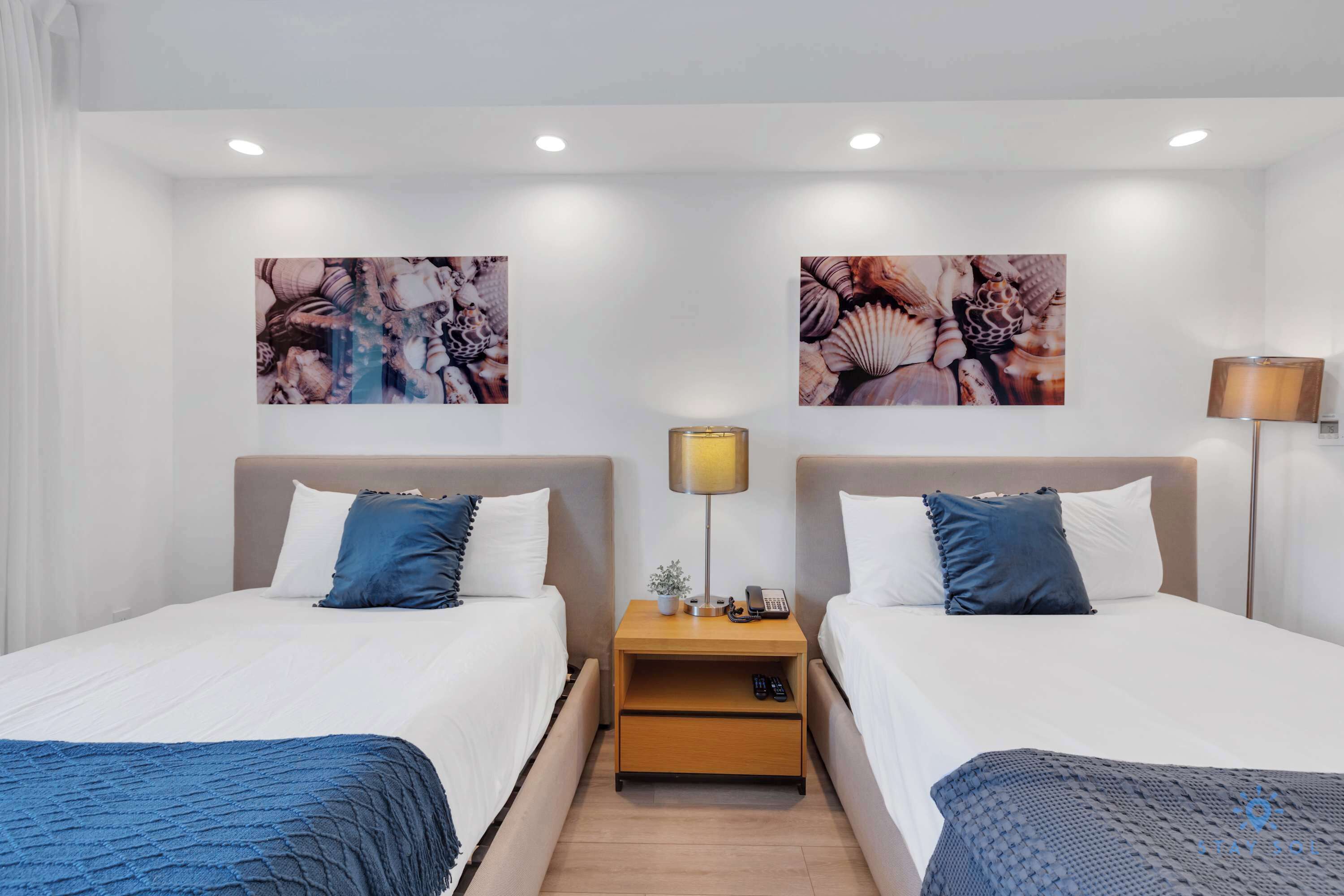 A hotel room with two beds, a nightstand with a lamp, and two wall-mounted artwork pieces.