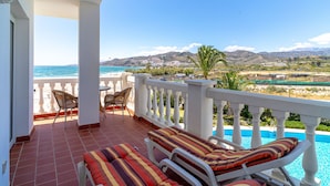 A balcony with a pool, lounge chairs, and a view of the sea and mountains.