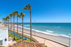 A beach view with palm trees, a tiled roof, and a row of blue lounge chairs. - Stella Maris Beachfront 1J by Casasol (Nerja)