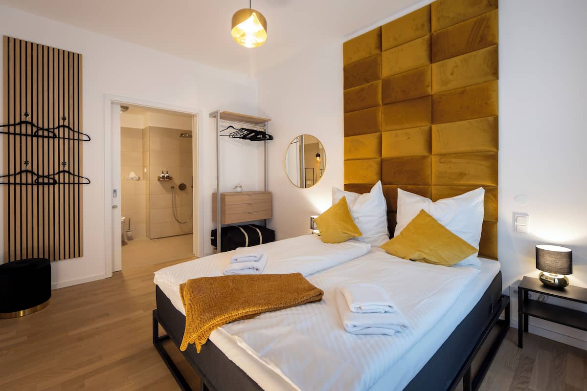 A hotel room with a large bed, a bedside table with a lamp, a wardrobe, a mirror, and a hanging coat rack.